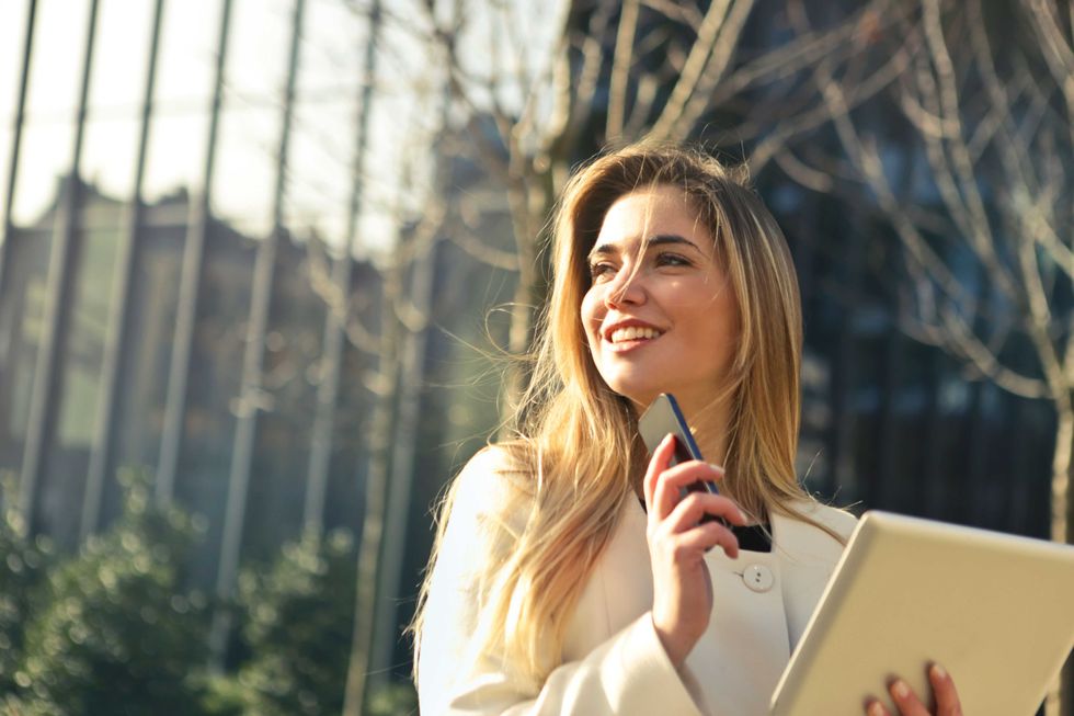 Smiling woman outdoors holding a phone and tablet, with trees in the background.