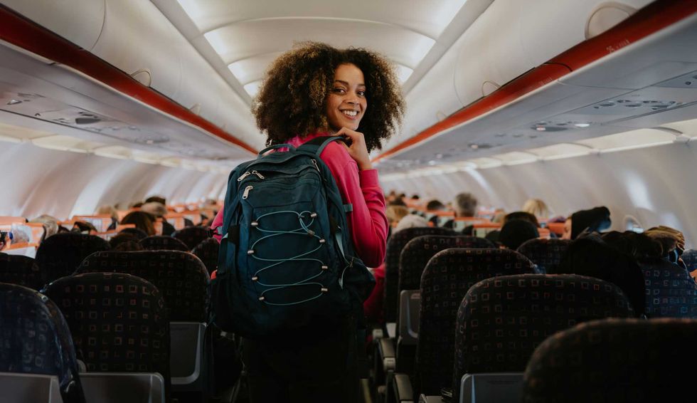 Smiling woman with a backpack stands in an airplane aisle, looking back toward the camera.