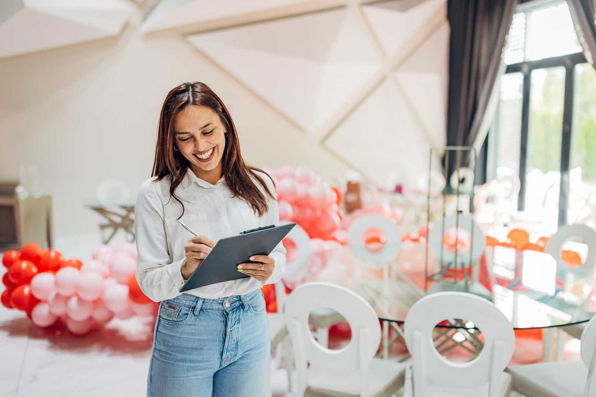 Smiling woman with clipboard stands in decorated room with pink and red balloons.