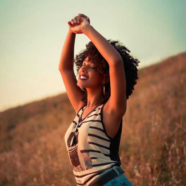 Smiling woman with curly hair stretches arms up in a field during sunset.