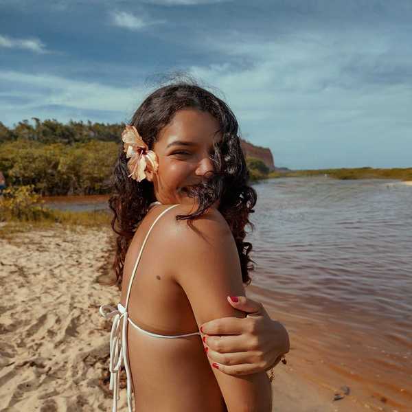 Smiling woman with flower in hair, standing by a river on a sandy beach.
