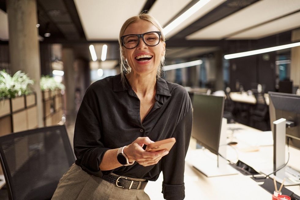 Smiling woman with glasses holding a phone in a modern office setting.
