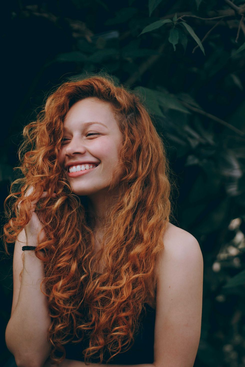 Smiling woman with long, curly red hair against a leafy background.