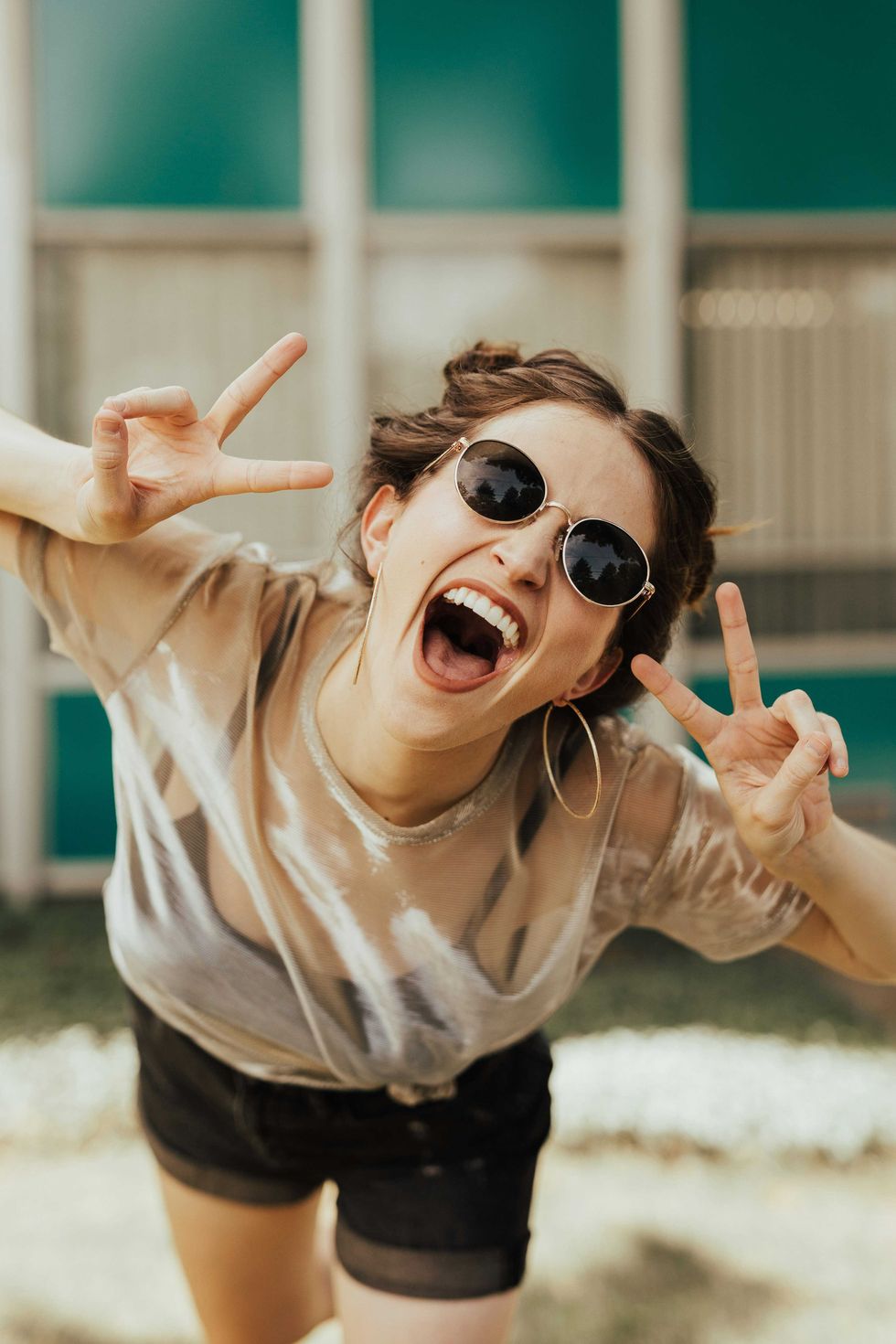 Smiling woman with peace signs, sunglasses, and hoop earrings, outdoors.