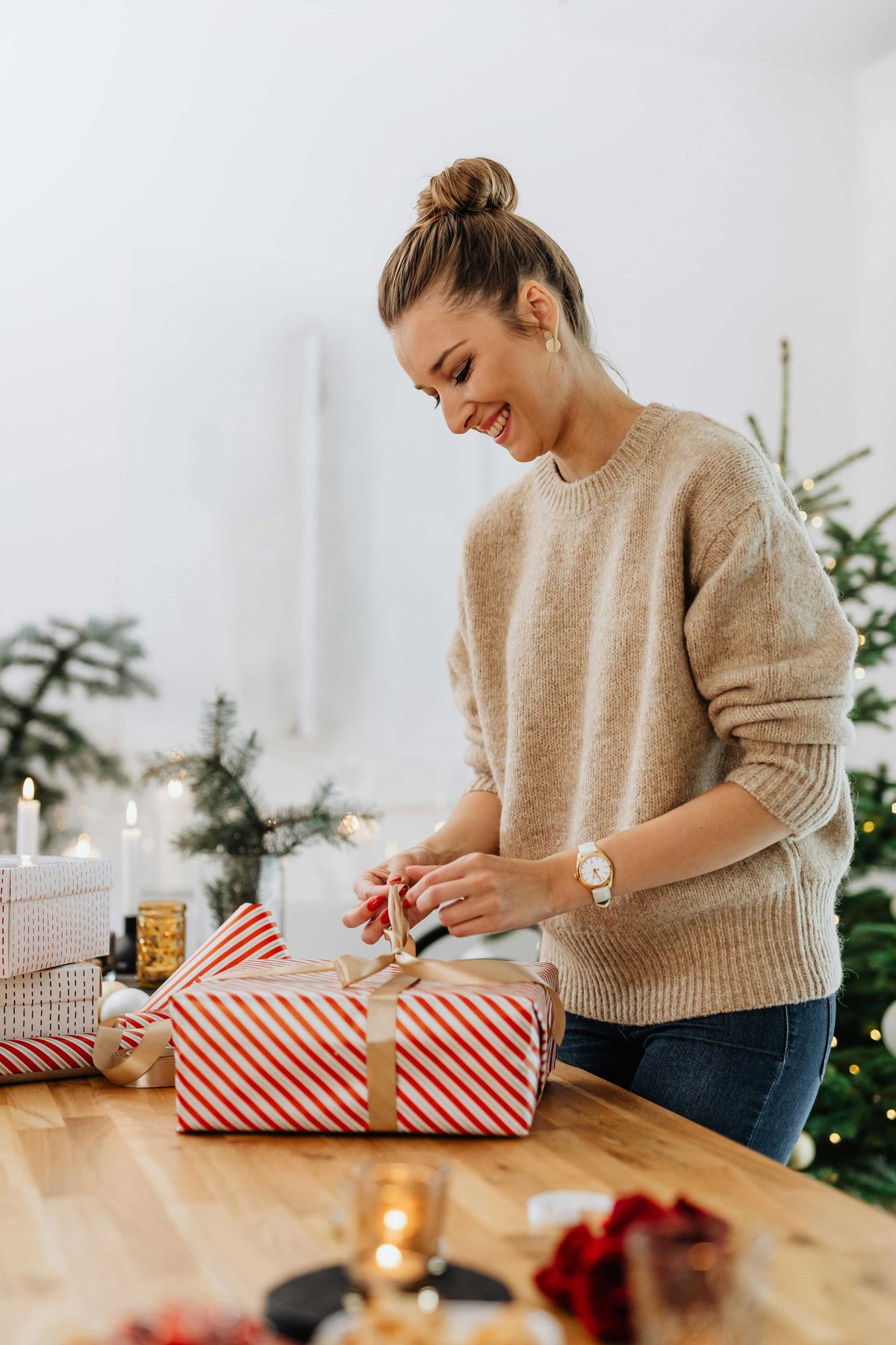 Smiling woman wrapping gifts at a wooden table, festive decor in background.