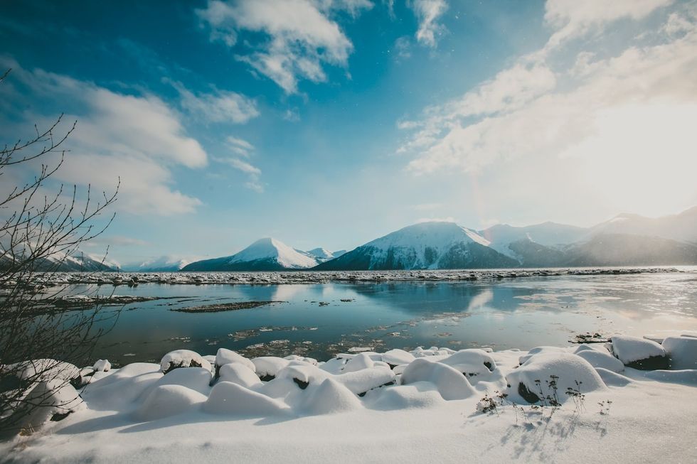 Snow-covered landscape with a calm lake and distant snowy mountains under a blue sky.
