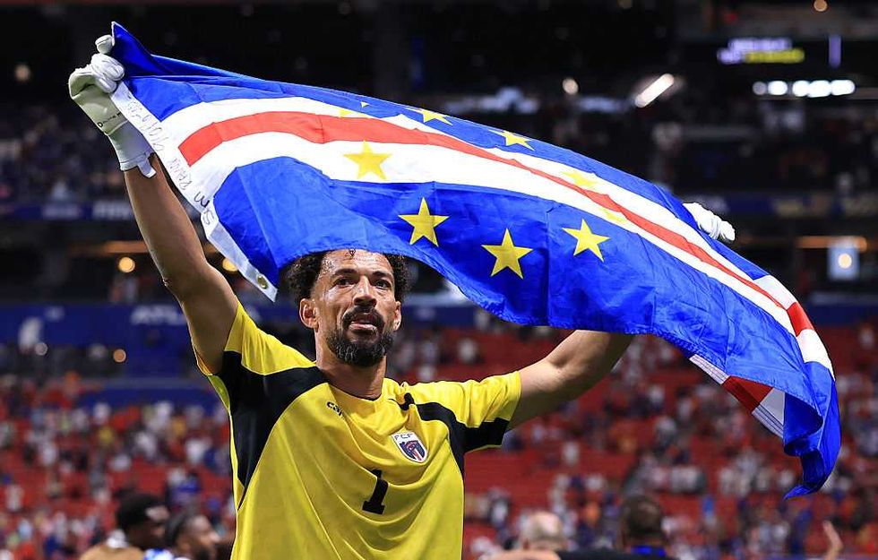 Soccer player in yellow jersey holds Cape Verde flag triumphantly on the field.