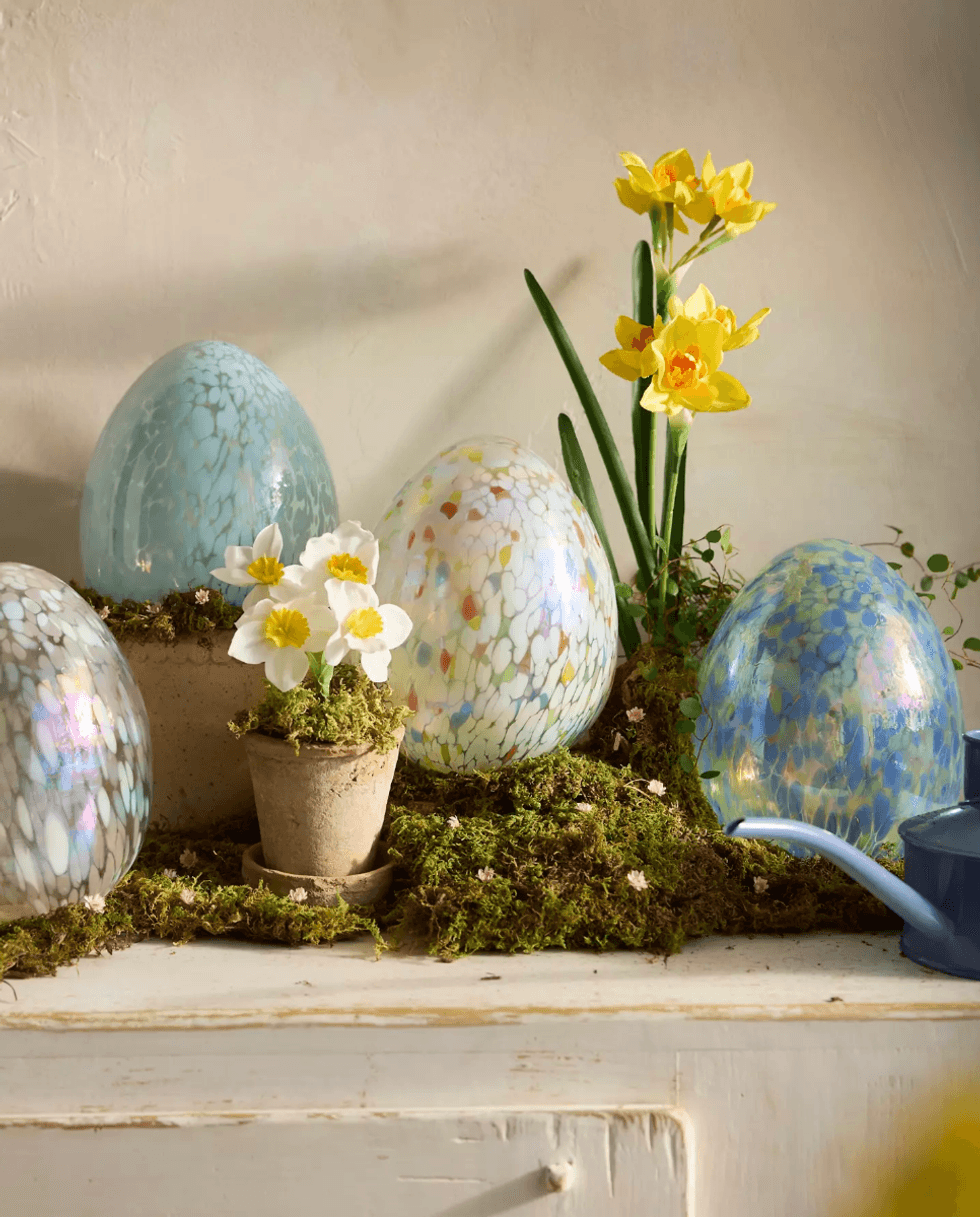 Speckled eggs, daffodils, and primroses on mossy surface with blue watering can.