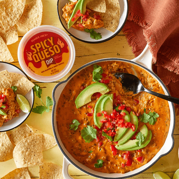 Spicy queso dip with avocado, tortilla chips, and lime on a rustic yellow table.