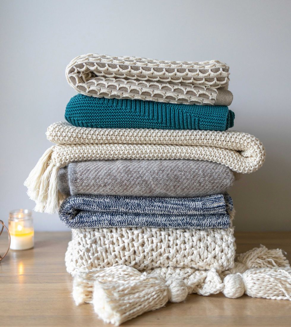 Stack of cozy, folded blankets in various textures on a wooden surface with a lit candle nearby.