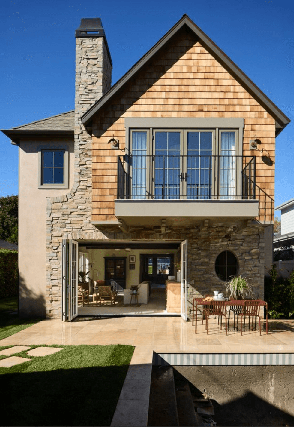 Stone house with balcony, large windows, open patio, and outdoor dining set.