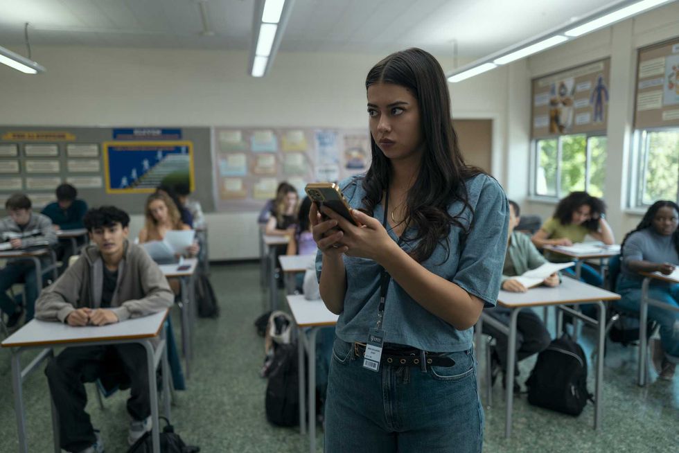 Student in a classroom intently looking at their phone, others seated at desks studying.