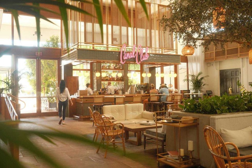 Stylish indoor cafe with a neon "Canal Club" sign, wooden furniture, and lush plants.