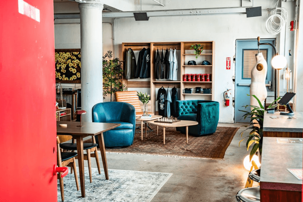 Stylish lounge with chairs, mannequins, plants, and clothing shelves, viewed from a red door.