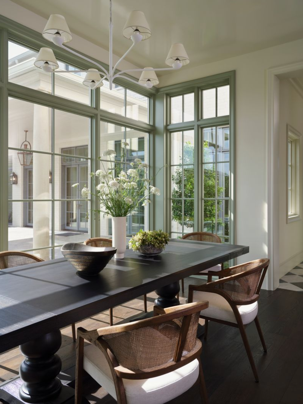 Sunlit dining room with a dark table, chairs, and large windows.