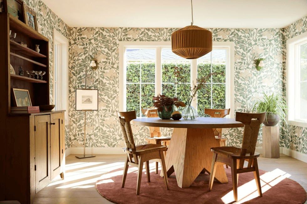 Sunlit dining room with floral wallpaper, wooden table, and chairs.