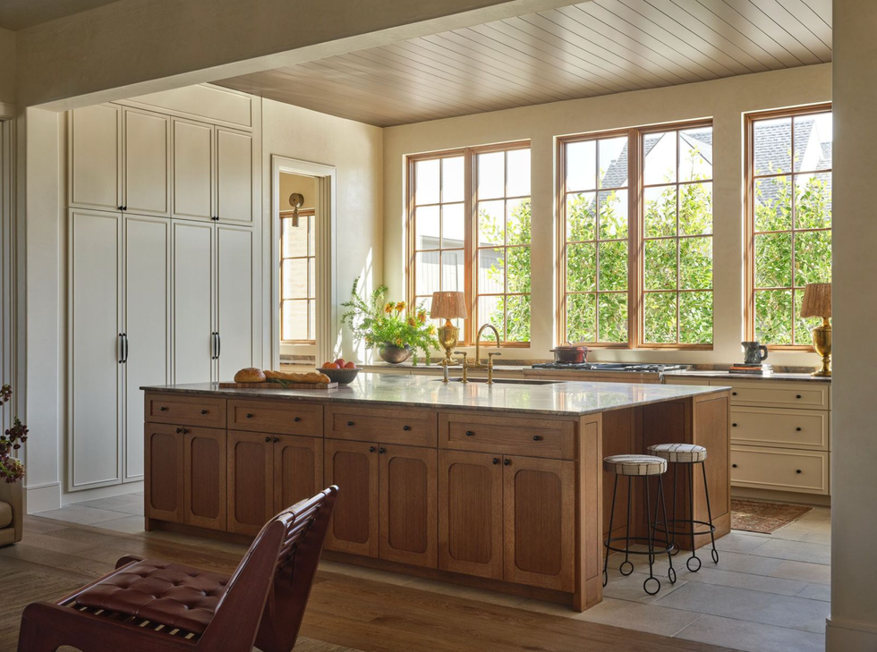 Sunlit kitchen with wooden island, white cabinets, and large windows overlooking greenery.