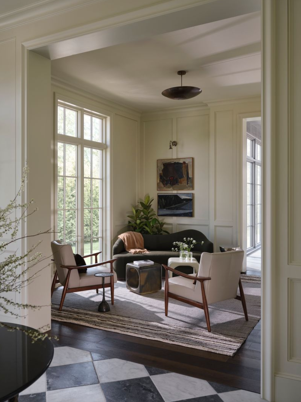 Sunlit living room with armchairs, sofa, and wall art by large windows.