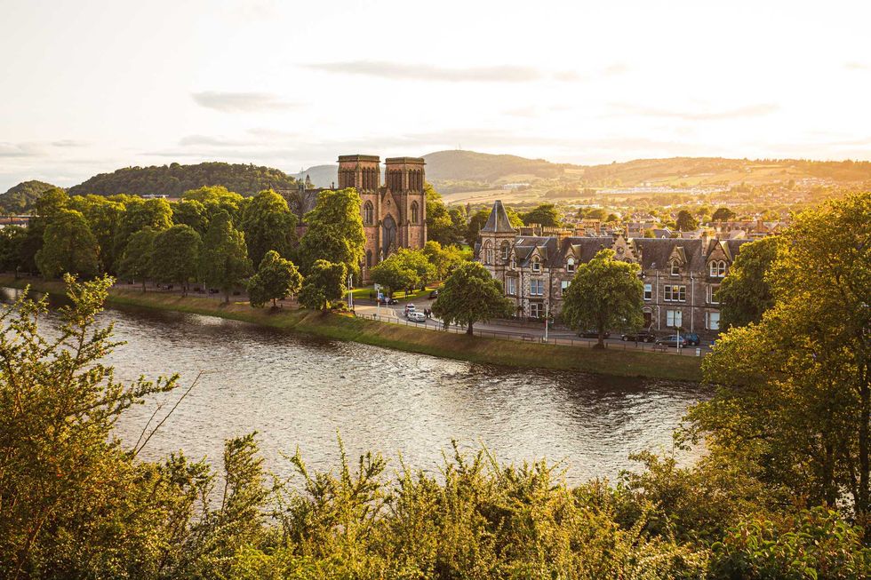 Sunlit riverside cityscape with cathedral, trees, and hills in the background.