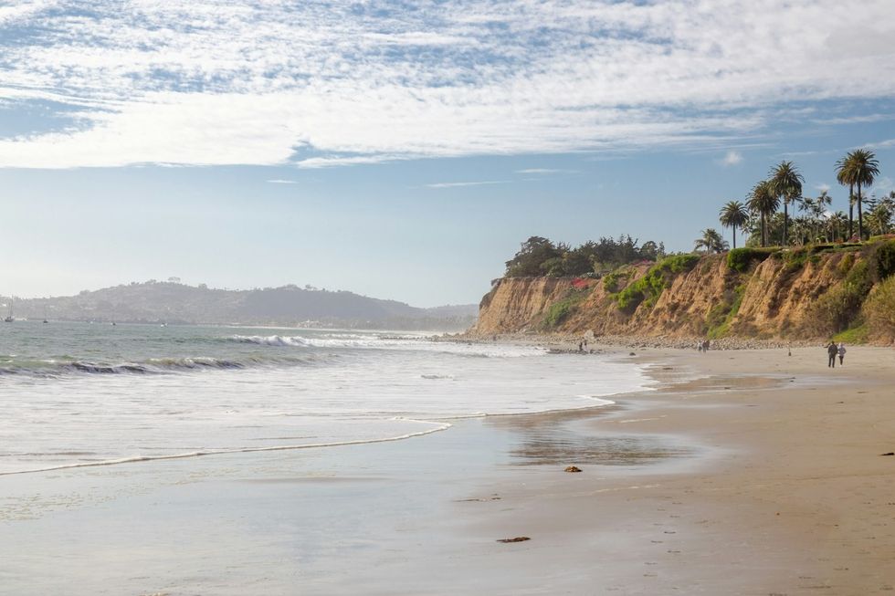 Sunny beach with cliffs, palm trees, and gentle waves under a partly cloudy sky.
