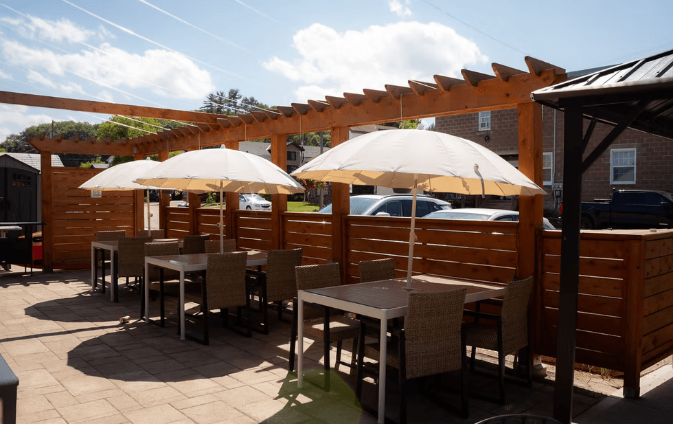 Sunny outdoor patio with tables, chairs, and white umbrellas under a wooden pergola.