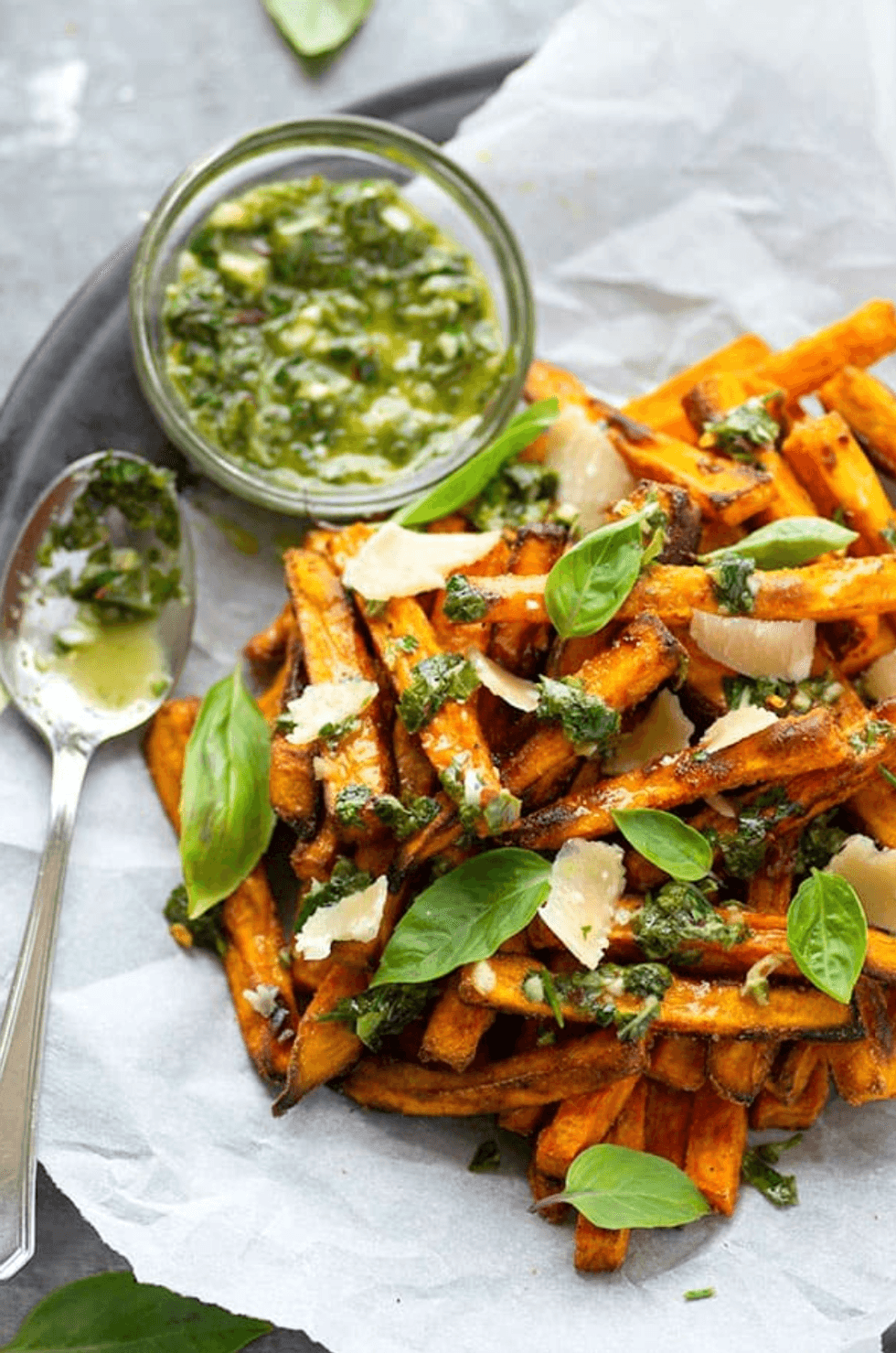 Sweet potato fries topped with pesto, garnished with basil and parmesan, beside a jar of pesto.
