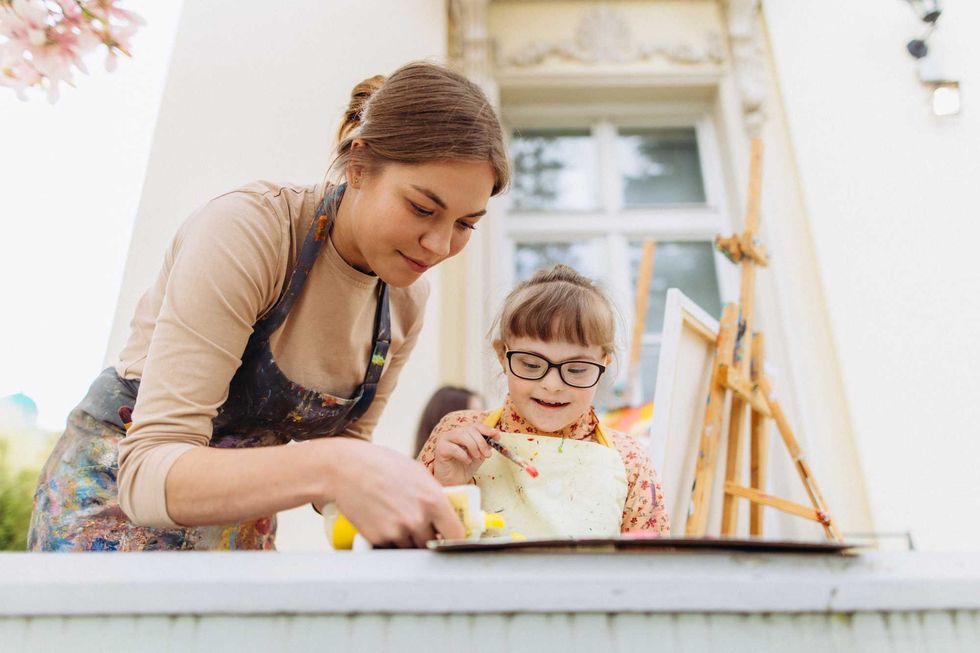 Teacher and child painting together outdoors, both wearing aprons.