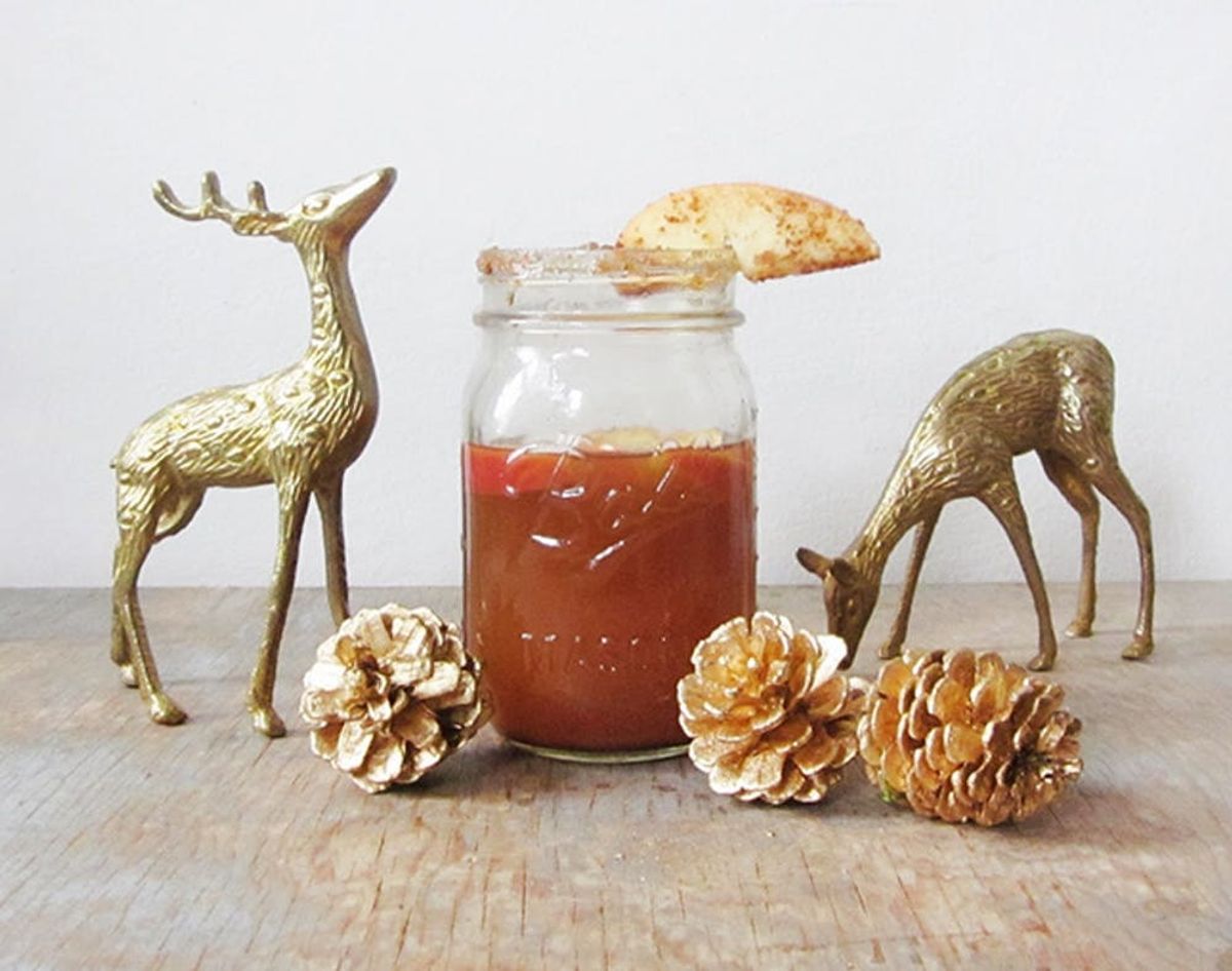 This list of 14 hot drinks includes this warm apple cider in a mason jar on a worn table with gold dipped pine cones and small gold deer figurines.