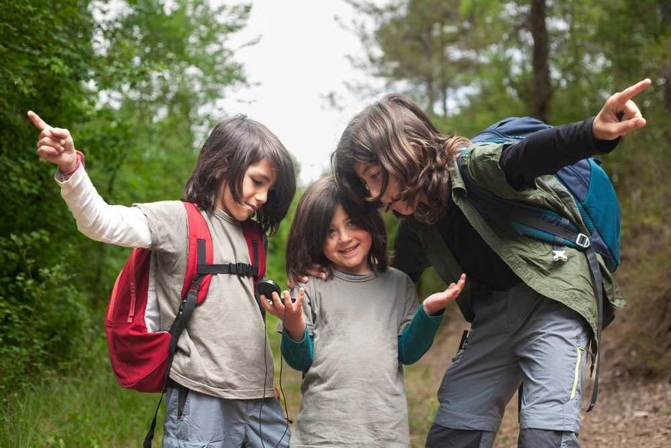 Three children with backpacks exploring a forest trail, pointing and smiling.