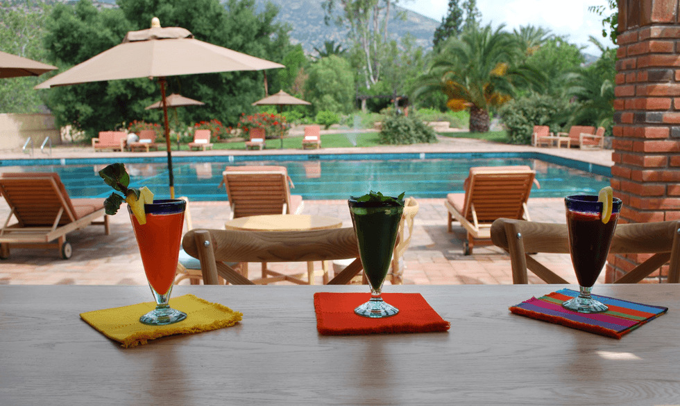 Three colorful drinks by a poolside with lounge chairs and umbrellas in the background.