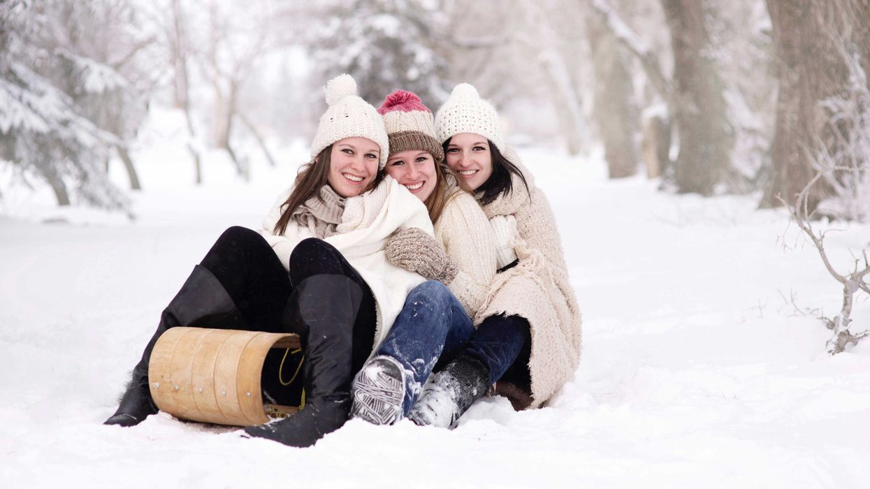Three friends in winter clothes sitting on a snowy path with a sled.