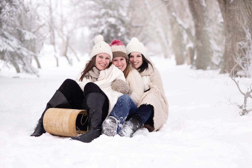 Three friends in winter clothes sitting on a snowy path with a sled.