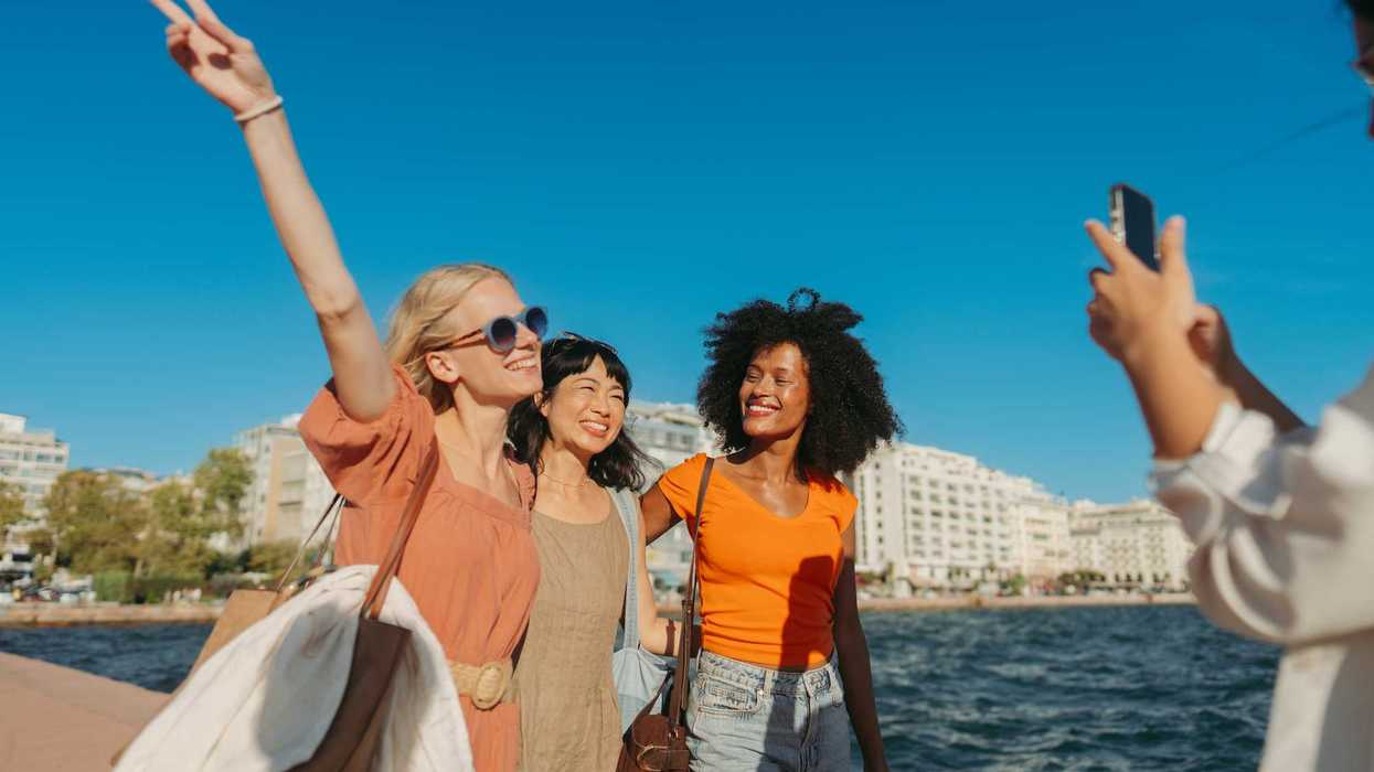 Three friends posing by the waterfront, one flashing a peace sign, with cityscape in background.