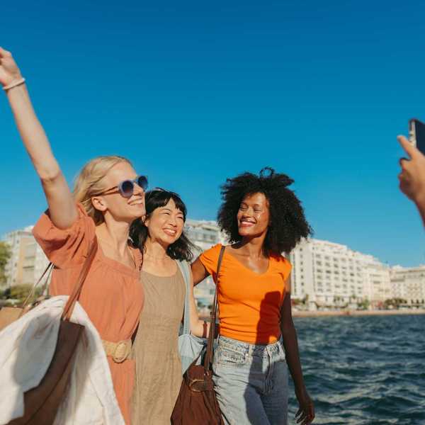 Three friends posing by the waterfront, one flashing a peace sign, with cityscape in background.