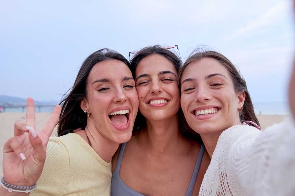 Three friends smiling widely, taking a selfie on a beach.