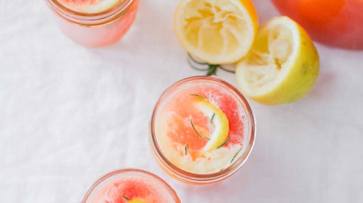 Three glasses of pink lemonade with lemon slices and rosemary on a white cloth.