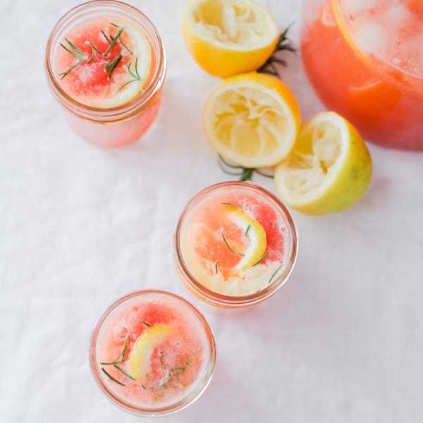 Three glasses of pink lemonade with lemon slices and rosemary on a white cloth.