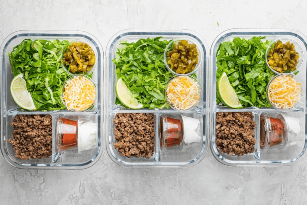 Three meal prep containers with taco ingredients, including beef, lettuce, cheese, and lime.