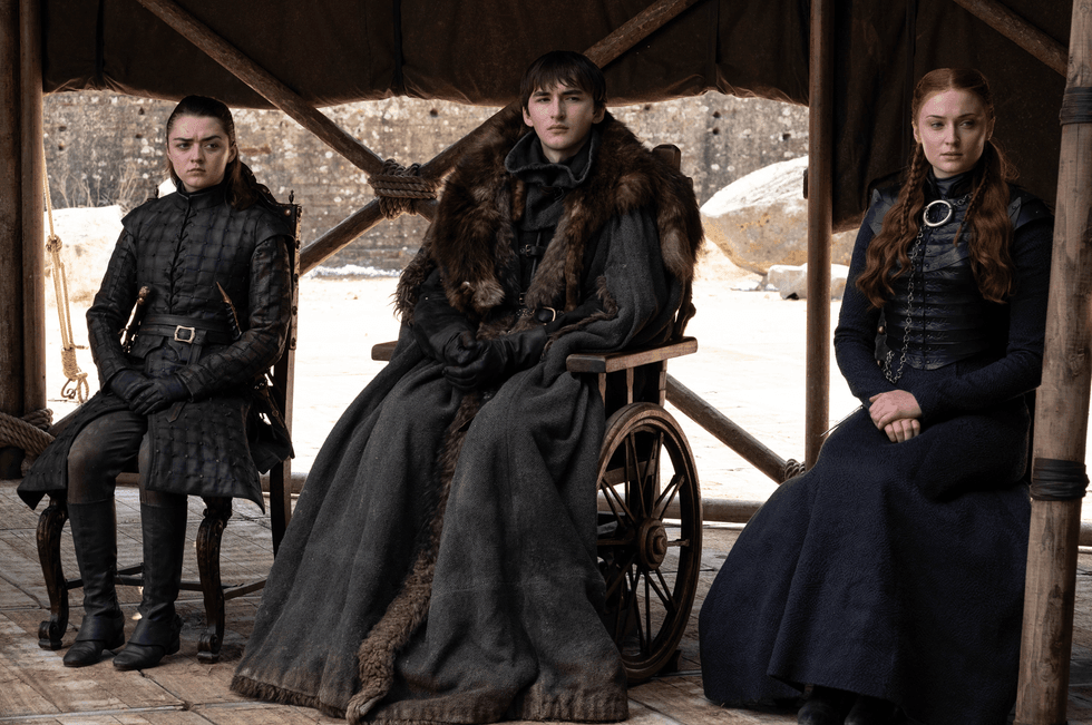 Three medieval-dressed individuals sit under a tent, one in a wheelchair, serious expressions.