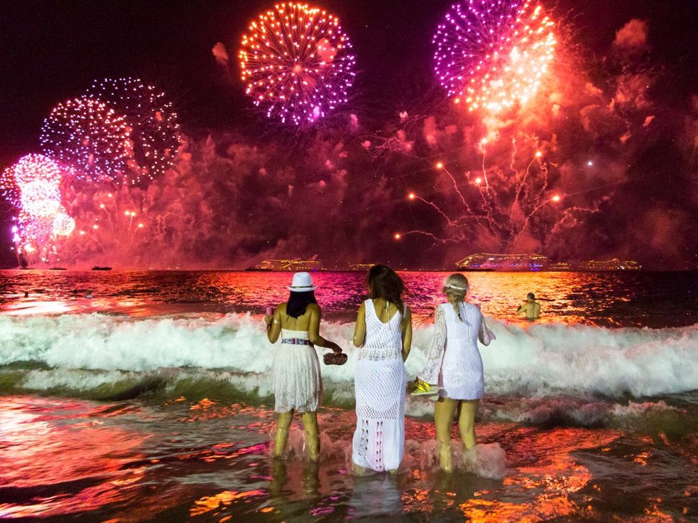 Three people on a beach watching vibrant fireworks over the ocean at night.