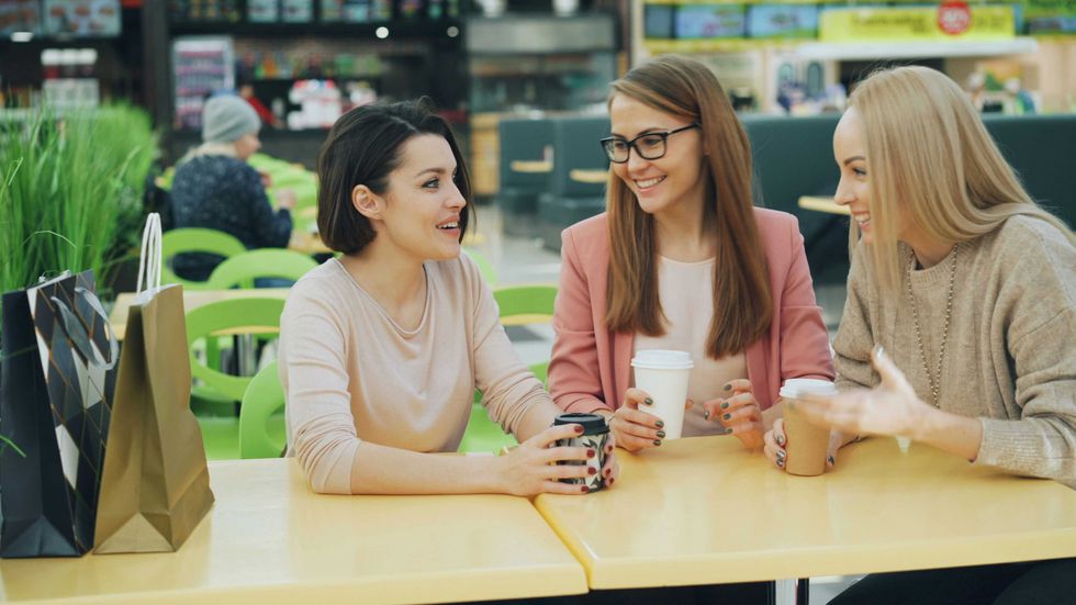 Three women chatting over coffee at a table with shopping bags nearby.