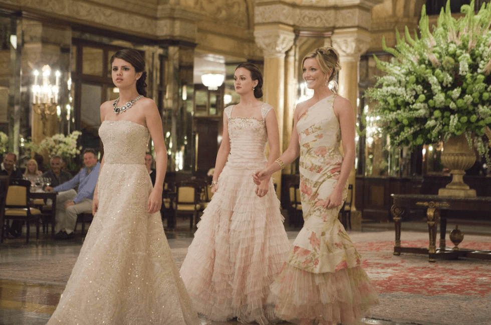Three women in elegant gowns walking through a luxurious, ornate room.