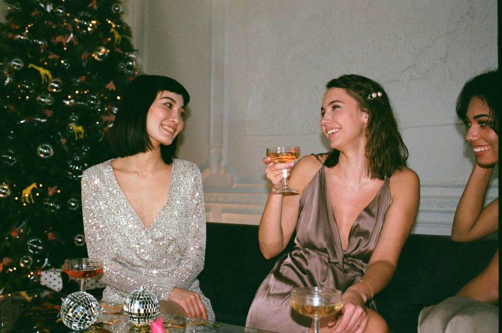 Three women laughing, holding drinks by a Christmas tree.