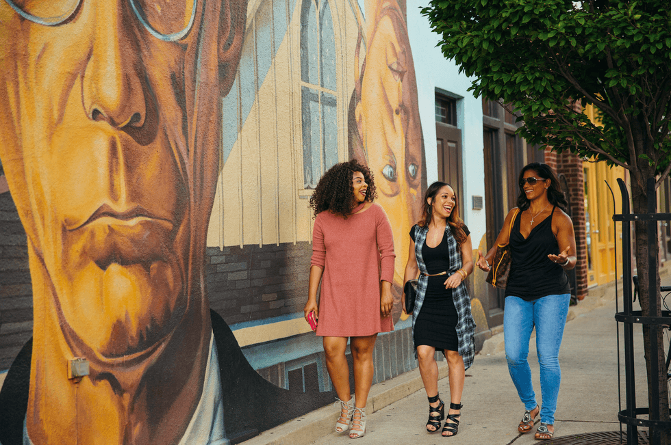 Three women walk and chat beside a colorful mural on a city sidewalk.
