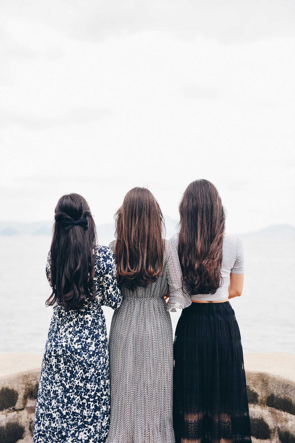 Three women with long hair standing by the sea, wearing patterned dresses.