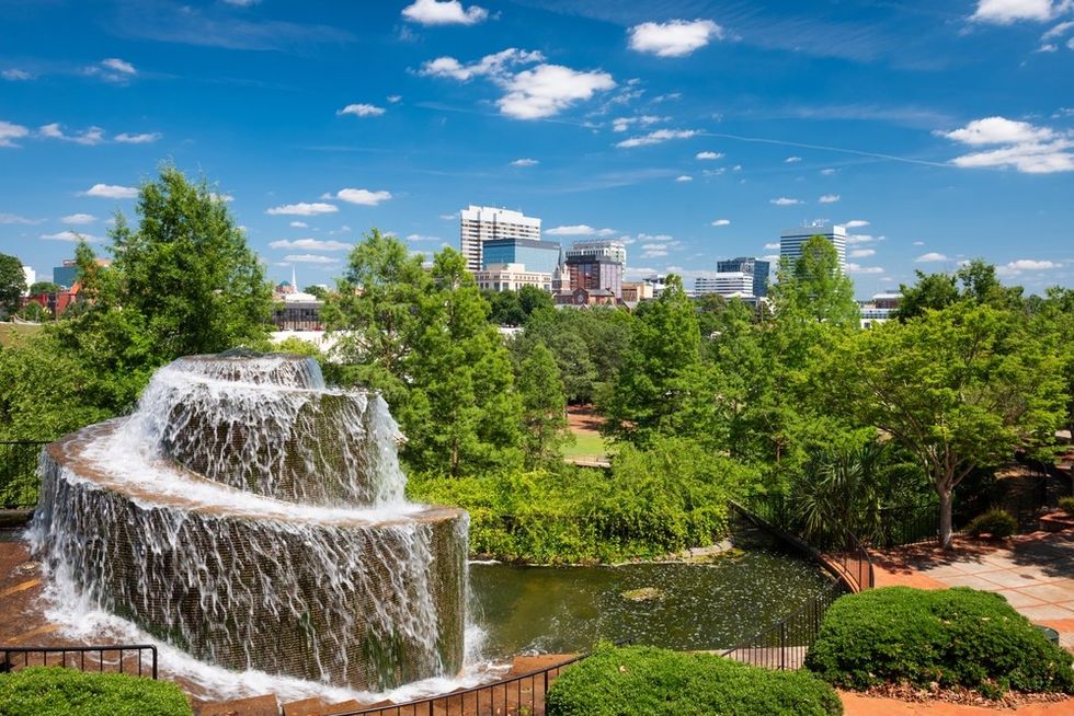 Tiered waterfall with city skyline and green trees under a bright blue sky.
