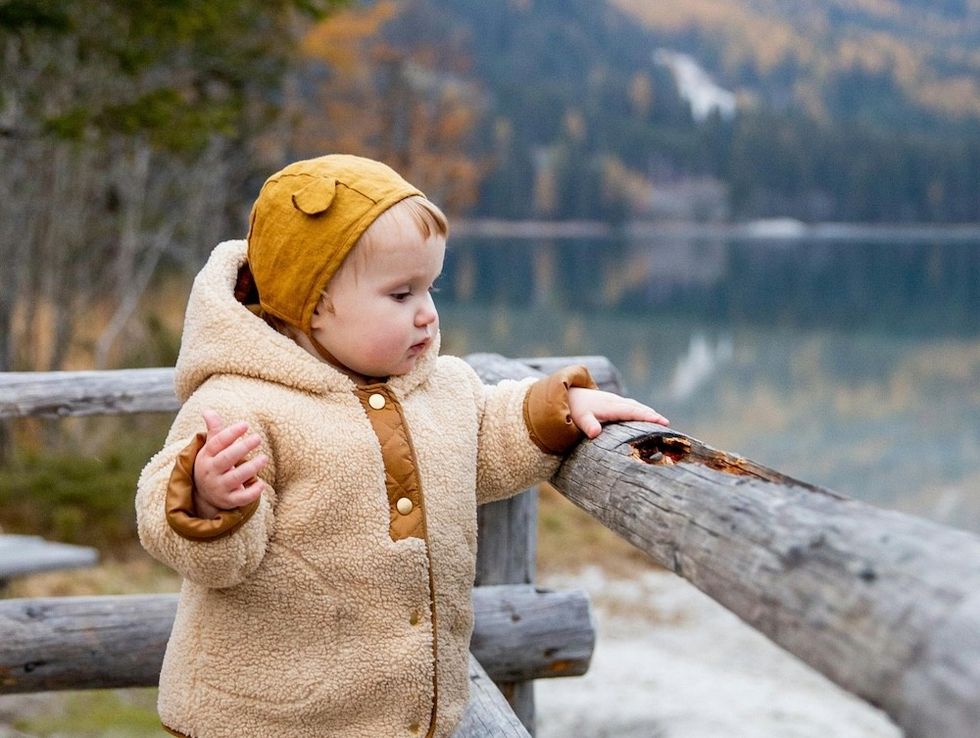 Toddler in cozy coat and hat leans on rustic fence by a serene lake with autumn trees.