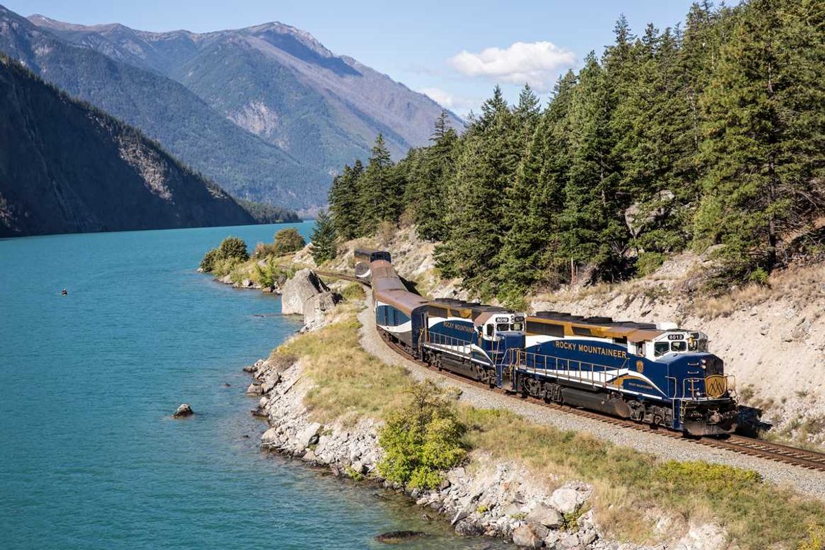 Train by turquoise lake, mountains, and forest in scenic landscape.