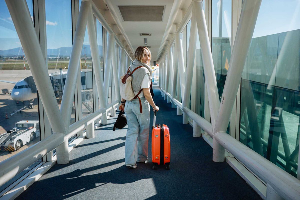 Traveler with orange suitcase walking through an airport jet bridge, smiling back.