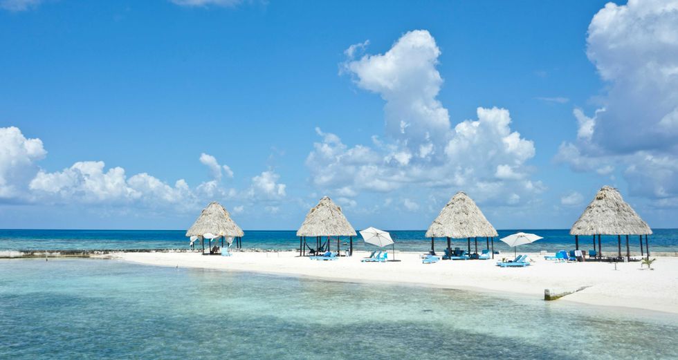 Tropical beach with huts, umbrellas, and clear blue water under a partly cloudy sky.